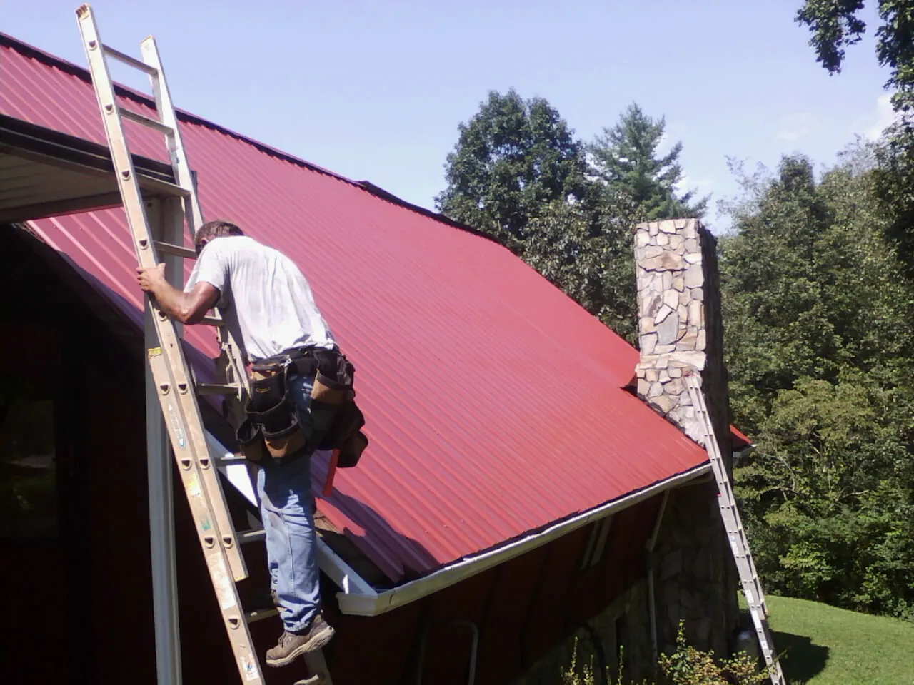 Roofer on a ladder beside a red metal roof and stone chimney — Soffit & Fascia Repair by Roofing Brothers in Alamo